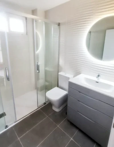 Un baño moderno con ducha de mampara de vidrio, lavabo blanco sobre mueble gris y un espejo redondo con luz LED integrada.