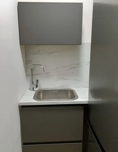 Cocina compacta con muebles grises y fregadero de acero inoxidable sobre una encimera de mármol blanco.