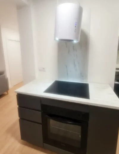 Vista de una cocina con campana extractora blanca suspendida sobre una placa de vitrocerámica, muebles oscuros y una encimera blanca de mármol.