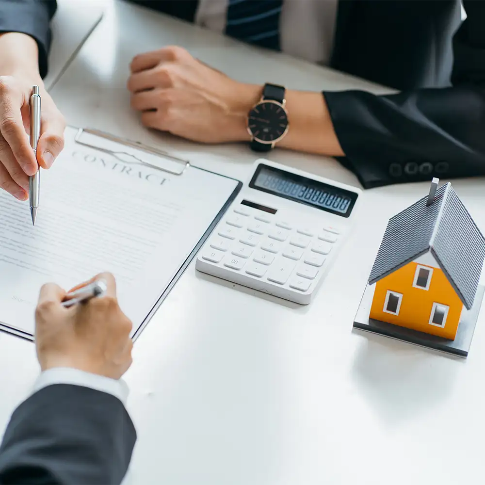 Personas firmando un contrato con una calculadora y una casa en miniatura en la mesa, representando un acuerdo de alquiler o compra de vivienda.
