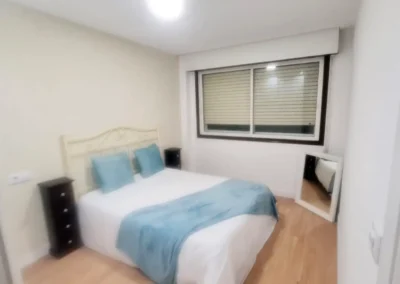 Dormitorio con paredes recién pintadas en tono beige claro, cama de matrimonio con cojines y mantas en tonos celeste y blanco.