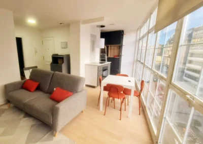 Comedor y salón con paredes pintadas en blanco, decorado con sofá gris y cojines rojos, grandes ventanales que permiten la entrada de luz natural.