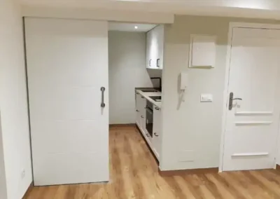 Vista de un salón con suelo de madera y una cocina pequeña integrada, separada por una puerta corredera blanca